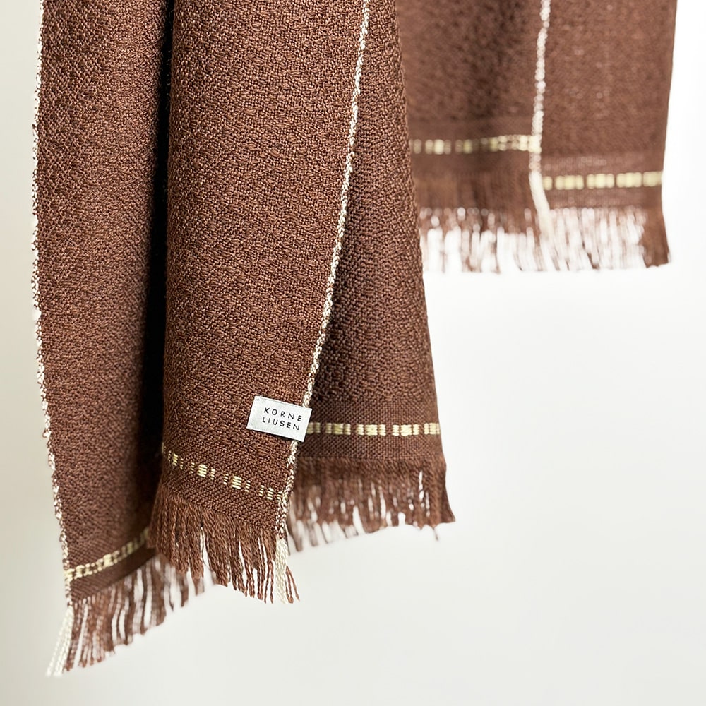 Ekslusivt håndvævet brunt tørklæde i merinould. Det er vævet brunt i brun ti et smukt mønster med lyse kanter der giver tørklædet karakter.